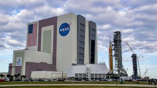 A bit orange rocket rolls out of a giant white building, with an extra launch tower in the background.