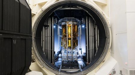 a tall cylindrical spacecraft wrapped in gold foil can be seen through a round door in a large metal chamber