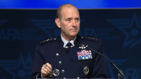 A man in a military uniform stands in front of a dark background. 